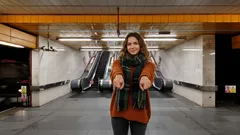 Žena ve stanic metra stojí před eskalátory a ukazuje oběma rukama dopředu.